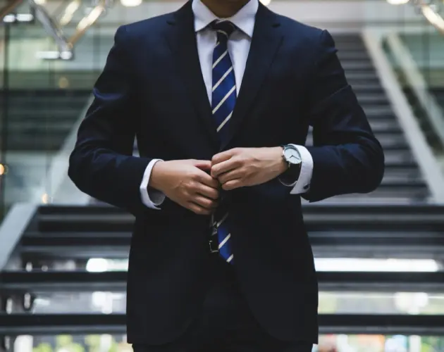 Hombre vestido con traje negro y corbata azul, ajustando su chaqueta mientras lleva un reloj elegante en la muñeca.